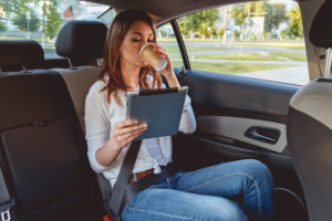 Young, beautiful woman sitting in the back seat of the car with a tablet in hand and drinking coffee